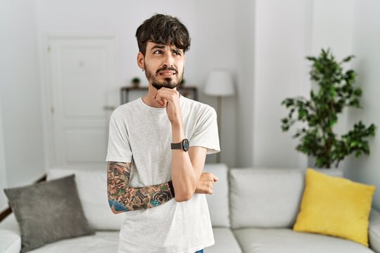 Hispanic man with beard at the living room at home thinking worried about a question, concerned and nervous with hand on chin