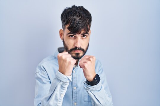 Young hispanic man with beard standing over blue background ready to fight with fist defense gesture, angry and upset face, afraid of problem