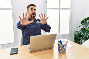 Young hispanic man with beard working at the office with laptop afraid and terrified with fear expression stop gesture with hands, shouting in shock. panic concept.