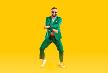 Fototapeta na wymiar Cheerful and stylish man is having fun dancing in Gangnam style at St. Patrick's Day party. Young man in trendy youth green suit and sunglasses is dancing and laughing on orange background. Banner.