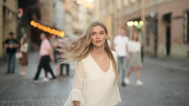 Attractive fashion blonde model in a white dress with flying hair. Young beautiful woman in a white dress spinning in the center of a city. Beautiful girl woman shakes hair. Slow motion