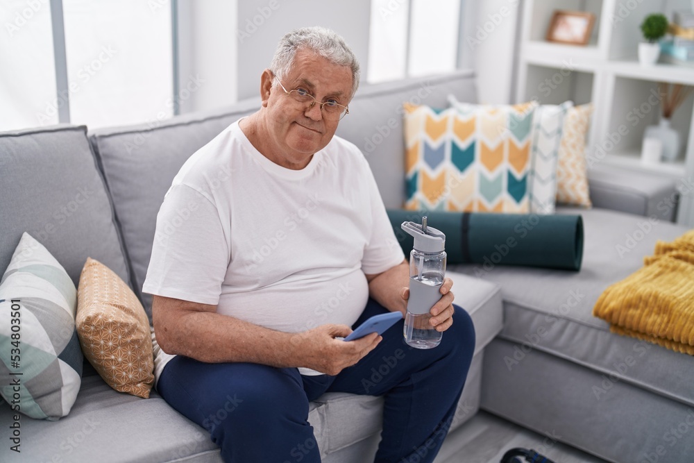 Sticker Middle age grey-haired man using smartphone holding bottle of water at home