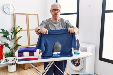 Senior caucasian man ironing holding burned iron shirt at laundry room looking at the camera...