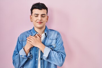 Non binary person standing over pink background smiling with hands on chest with closed eyes and grateful gesture on face. health concept.