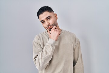 Young handsome man standing over isolated background looking confident at the camera smiling with crossed arms and hand raised on chin. thinking positive.