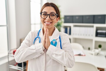 Young brunette doctor woman wearing stethoscope at the clinic praying with hands together asking for forgiveness smiling confident.