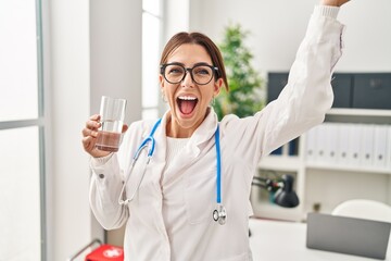 Young brunette doctor woman holding glass of water celebrating victory with happy smile and winner expression with raised hands