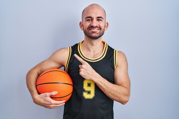 Obraz premium Young bald man with beard wearing basketball uniform holding ball cheerful with a smile on face pointing with hand and finger up to the side with happy and natural expression