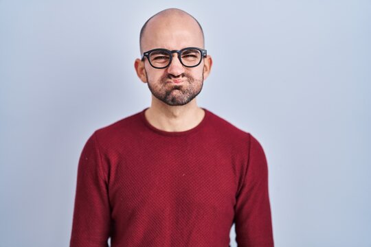 Young bald man with beard standing over white background wearing glasses puffing cheeks with funny face. mouth inflated with air, crazy expression.
