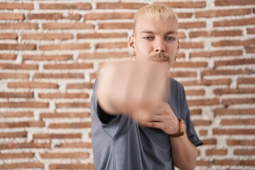Young caucasian man standing over bricks wall punching fist to fight, aggressive and angry attack, threat and violence