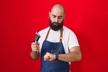 Young hispanic man with beard and tattoos wearing barber apron holding razor checking the time on...