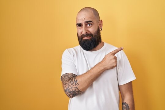 Young hispanic man with beard and tattoos standing over yellow background pointing aside worried and nervous with forefinger, concerned and surprised expression