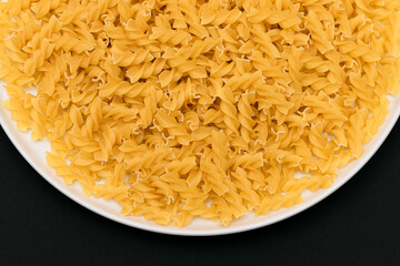 Uncooked Fusilli Pasta Lying on White Plate on Black Background. Raw and Dry Macaroni. Unhealthy and Fat Food - Top View