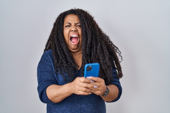 Plus Size Hispanic Woman Using Smartphone Typing Message Angry And Mad Screaming Frustrated And Furious, Shouting With Anger. Rage And Aggressive Concept.