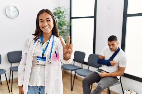 Young asian doctor woman at waiting room with a man with a broken arm smiling looking to the camera showing fingers doing victory sign. number two.