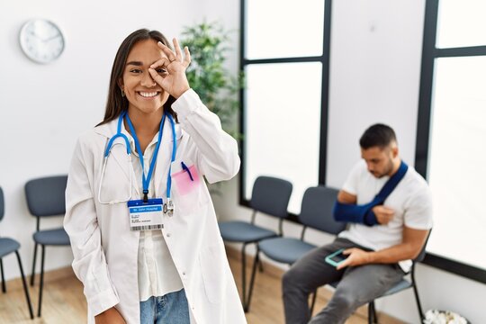 Young asian doctor woman at waiting room with a man with a broken arm doing ok gesture with hand smiling, eye looking through fingers with happy face.