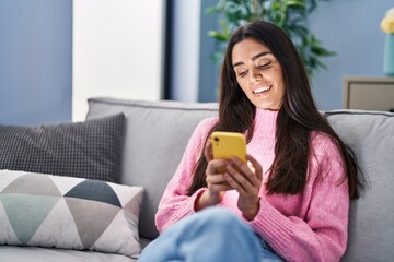 Obraz na płótnie Canvas Young hispanic woman using smartphone sitting on sofa at home