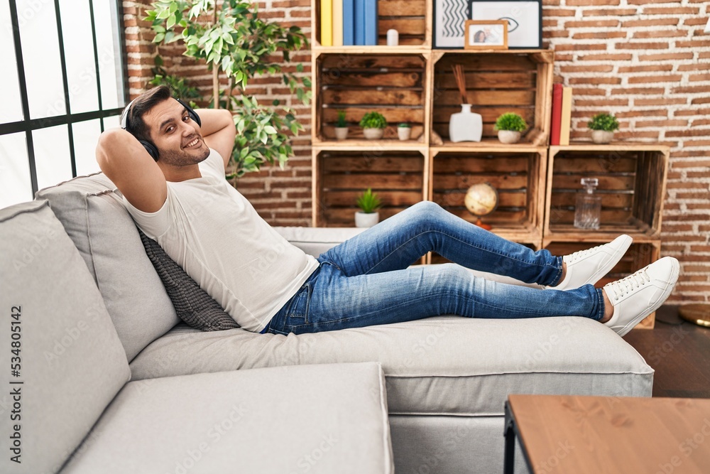 Wall mural Young hispanic man relaxed with hands on head listening to music at home