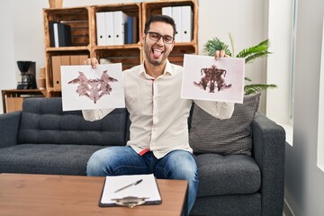 Young hispanic man with beard holding rorschach test sticking tongue out happy with funny expression.