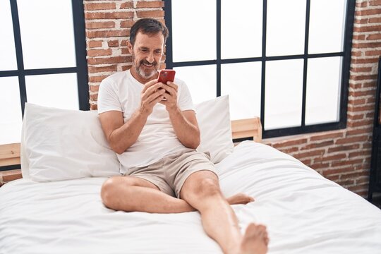 Middle age man using smartphone sitting on bed at bedroom - Powered by Adobe