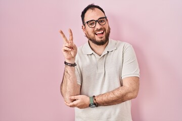 Plus size hispanic man with beard standing over pink background smiling with happy face winking at...