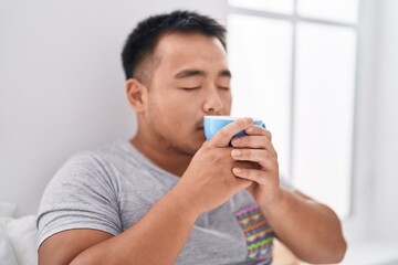 Young chinese man smelling cup of coffee sitting on bed at bedroom