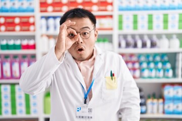 Chinese young man working at pharmacy drugstore doing ok gesture shocked with surprised face, eye...