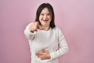 Woman with down syndrome standing over pink background laughing at you, pointing finger to the...