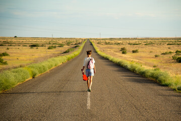 Obraz premium A girl walks along an empty road in the steppe. She has a ukulele in her hands and a backpack on her shoulders. It's summer and warm outside. Open road.