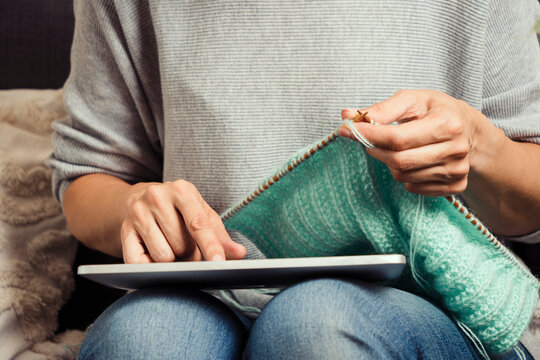 Woman holding digital tablet computer on her lap, looking at screen and knitting sweater. Woman learning knitting, watching video tutorial, online class lesson, follow knitting patterns