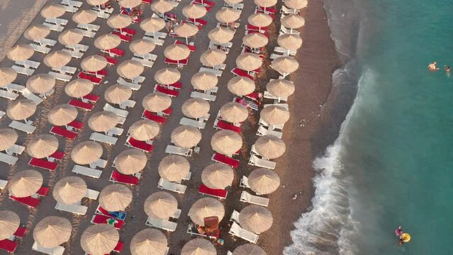 Beach with rows of thatch umbrellas - tiki straw parasols by the sea. Waves crashing on the sand (lapping at the shore). Aerial drone view on the coast at a summer vacation resort.