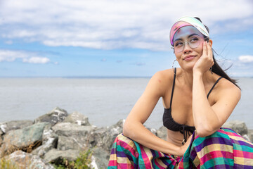 woman in bikini on the beach wearing a visor and colorful pants. Her hand is resting on her chin and she is looking up and thinking. 