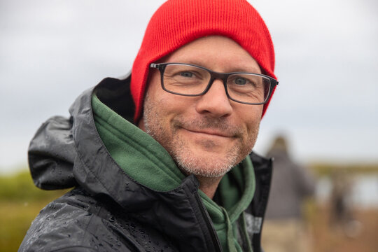 Close up face of handsome man wearing red beanie hat and glasses. He has beard stubble and a friendly smile. 