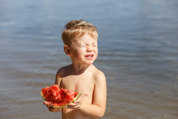 A blond boy 3-4 years old eats a piece of ripe red watermelon, looks at the bright sun, closes his eyes against the background of the river. On a sunny summer day - obrazy, fototapety, plakaty