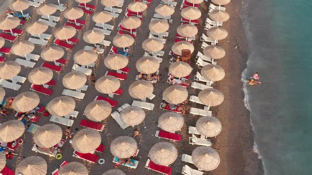 Beach with rows of thatch umbrellas - tiki straw parasols by the sea. Waves crashing on the sand (lapping at the shore). Aerial drone view on the coast at a summer vacation resort.