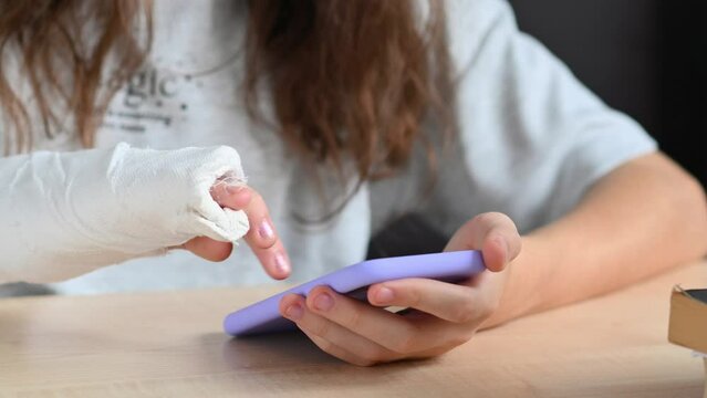 A teenage girl with a broken arm with a smartphone in her hands. Close-up of a girl with a bandage on her arm uses the phone. Selective focus on the fingers and bandage.