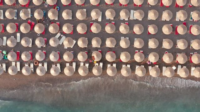 Beach with rows of thatch umbrellas - tiki straw parasols by the sea, waves crashing on the sand. Tourism on the Mediterranean. Aerial drone top down view on the coast at a summer vacation resort.
