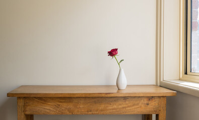 Single red rose in white vase on oak sidetable against beige wall next to window with sunlight