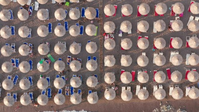 Beach with rows of thatch umbrellas - tiki straw parasols by the sea. Aerial drone top down view on the coast at a summer vacation resort.