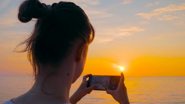 Slow motion: woman is holding smartphone and taking photo or shooting video of the Black Sea, warm sunset sky - close up, sun lens flares. Freedom, photography and summer concept