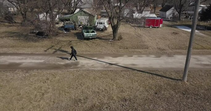 Aerial drone shot following a young high school aged teen boy wearing a backpack walking down a road during the winter on a cold day.