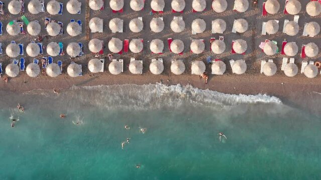 Beach with rows of thatch umbrellas - tiki straw parasols by the sea, waves crashing on the sand. Tourism on the Mediterranean. Aerial drone top down view on the coast at a summer vacation resort.