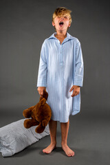 Cute little boy in a nightshirt yawning while holding his stuffed bear