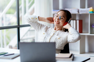 Cute young Asian woman wearing headphones listening to music happily on her laptop enjoying listening to work music during a relaxing break at work.