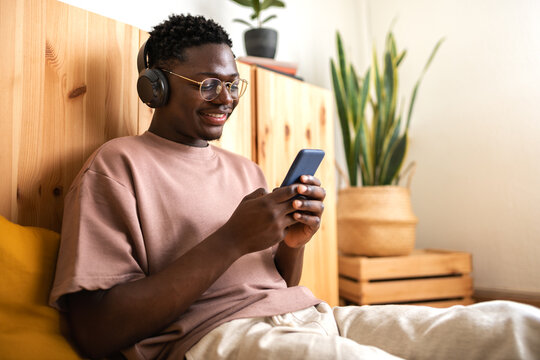 Young african american man with glasses relaxing at home listening to music with headphones while using mobile phone.