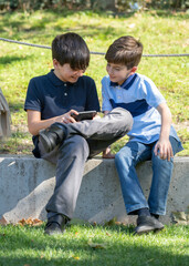 Cute two boys sitting on the park and play online games. One boy holding the smartphone. Young boys use the phone