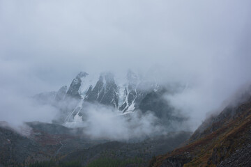 Dark atmospheric landscape with high snow mountains in dense fog in rainy weather. Large snowy mountain range in thick fog in dramatic overcast. Snow-covered rocky mountains in low clouds during rain.