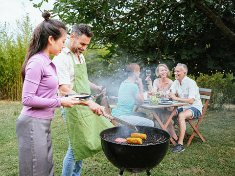 Couple cooking on a barbeque in countryside. At the back, his friends celebrating with alcohol drinks. Lifestyle concept. High quality photo