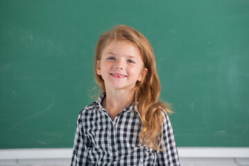 Close-up portrait of attractive small little cheerful girl sitting on table desktop in class room indoors. Little funny school girl face. Kids education and knowledge. Student kids.