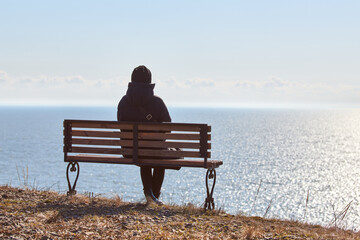 Single girl in a black jacket and hat sitting on bench at cliff at front of sea, peaceful and quiet...